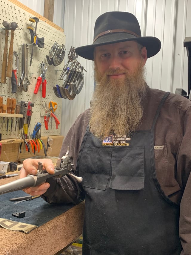 Ben in his shop, wearing his AGI Certified Gunsmith apron and holding a firearm he's working on.