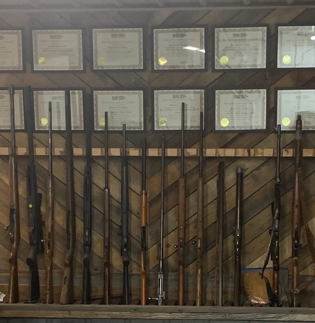 Rifles on a display rack below framed credentials on the shop wall.